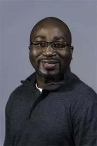 Male team member in dark hoodie and glasses smiling at camera against blue background