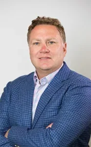 Male team member in blue checkered blazer and light pink shirt, smiling at camera with arms crossed