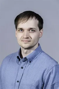 Male team member in light blue button-up shirt smiling at camera against gray background