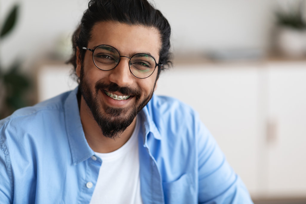 Closeup Portrait Of Positive Indian Guy With Dental Braces And Eyeglasses Smiling At Camera, Handsome Bearded Millennial Man With Brackets On Teeth Posing Indoors, Selective Focus With Copy Space