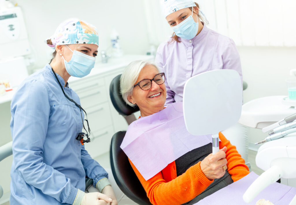 Happy senior woman holding a mirror and admiring her smile after a dental checkup with two dentists