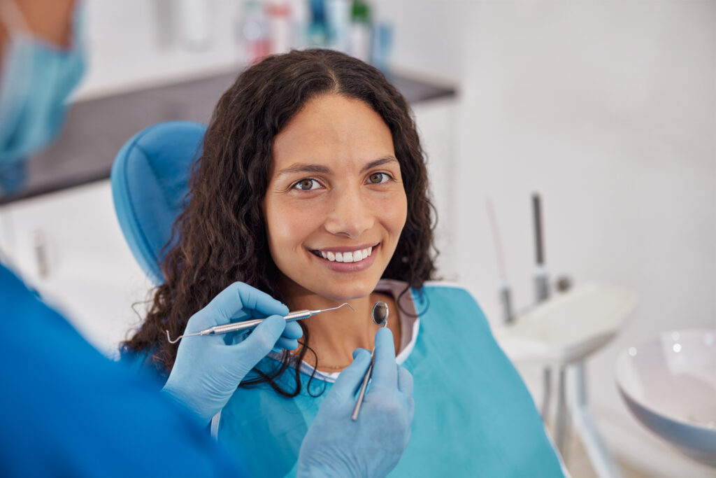 Happy young patient smiling while sitting in dental chair during oral hygiene. Young woman with curly hair wearing dental bib in bright clinic. Dentist holding tools near patient during consultation for whitening teeth treatment.