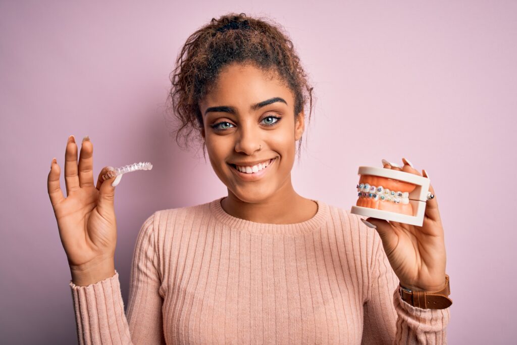 Young african american woman smiling happy holding professional orthodontic denture with metal braces and removable invisible aligner. Comparation of two dental straighten treatments