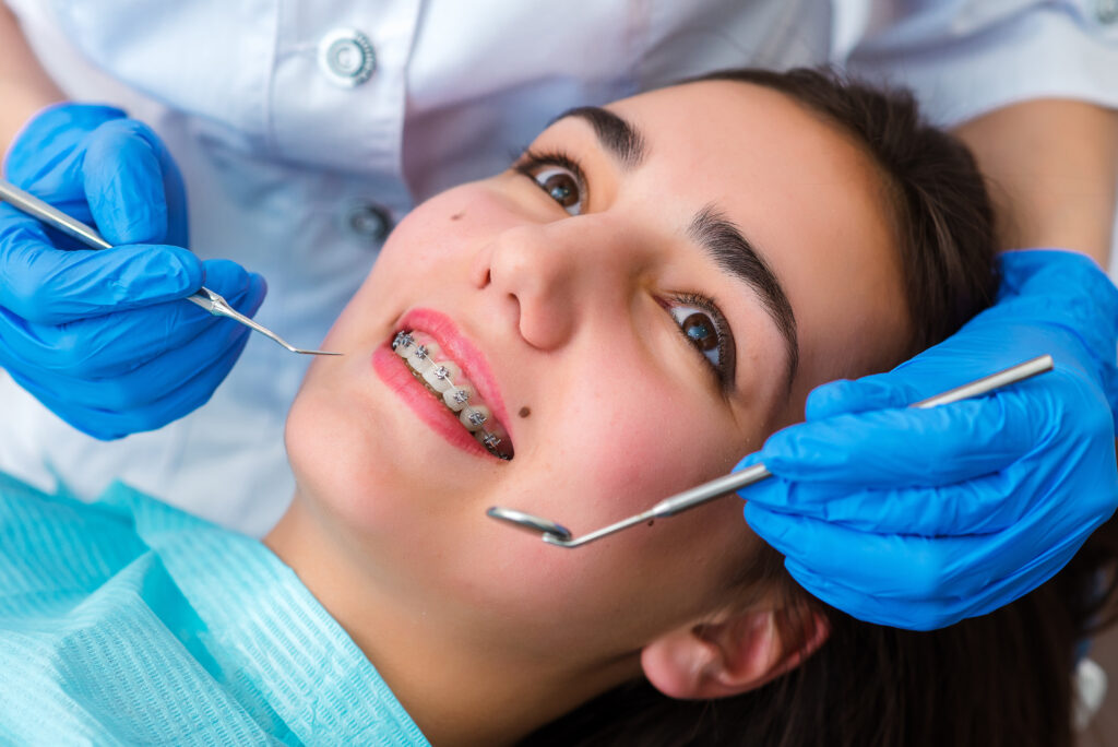 Close up of woman with brackets receiving dental braces treatment in clinic. Orthodontist using dental mirror and forceps while putting orthodontic braces on patient teeth. Concept of dentistry.