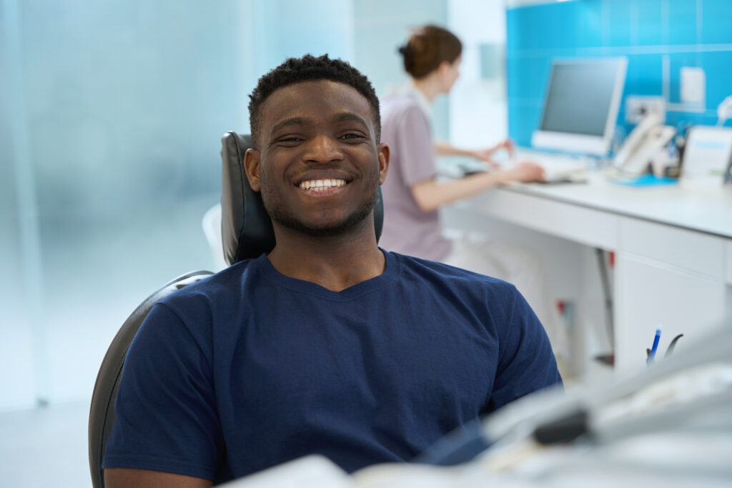 Male client sitting in dental chair with healthy and clean smile while stomatologist keeping medical records on computer