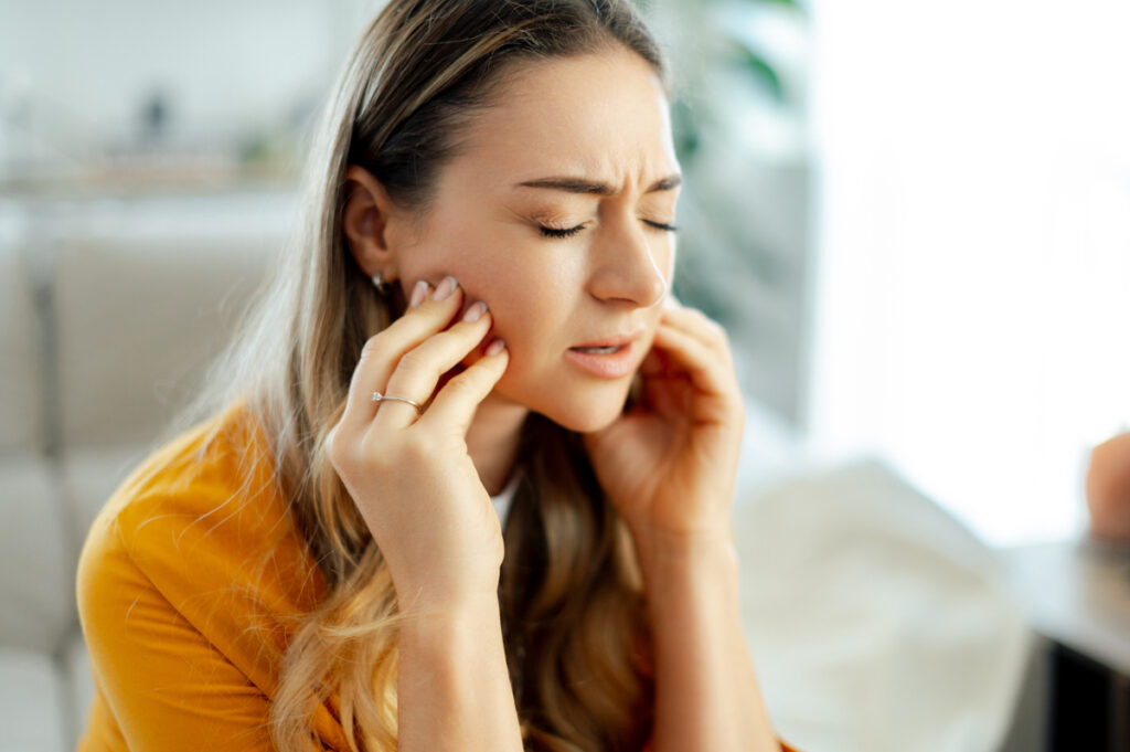 Tooth pain concept. Caucasian young woman in a yellow shirt sits on a couch at home with eyes closed, pressing her jaw with both hands and wincing, indicating tooth pain or discomfort inside mouth