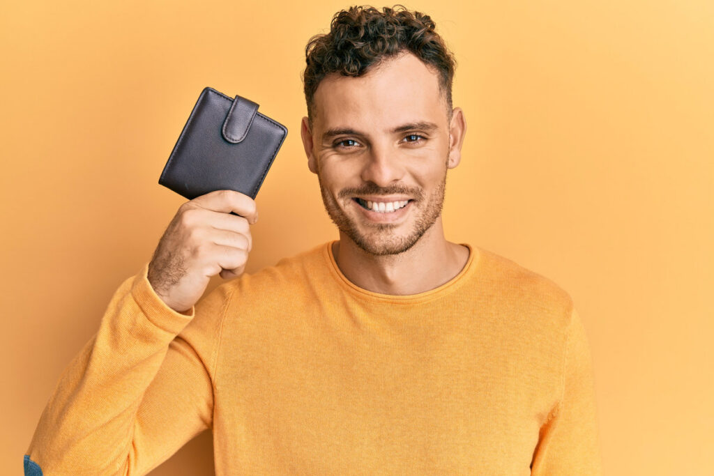 Young hispanic man holding leather wallet looking positive and happy standing and smiling with a confident smile showing teeth